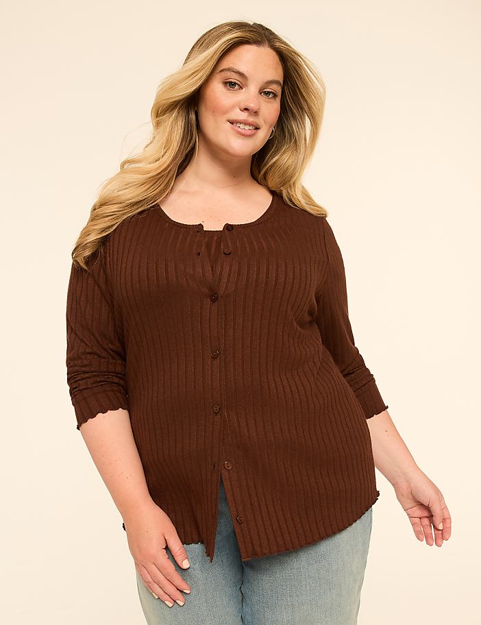 A woman in a brown cardigan and jeans stands against a plain, light-colored background.