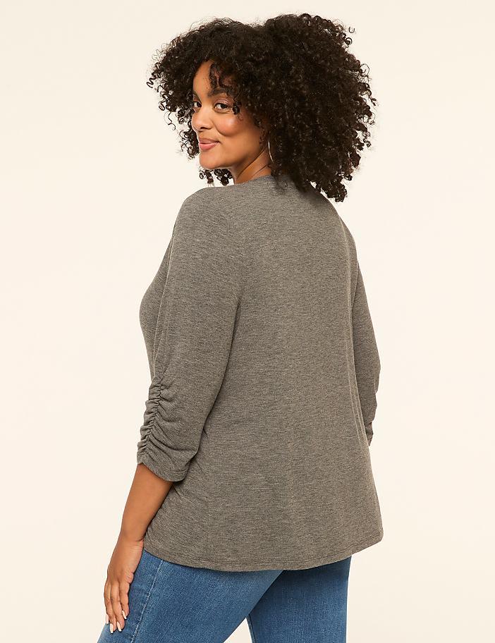 A woman in a grey cardigan and blue jeans stands against a plain cream background.