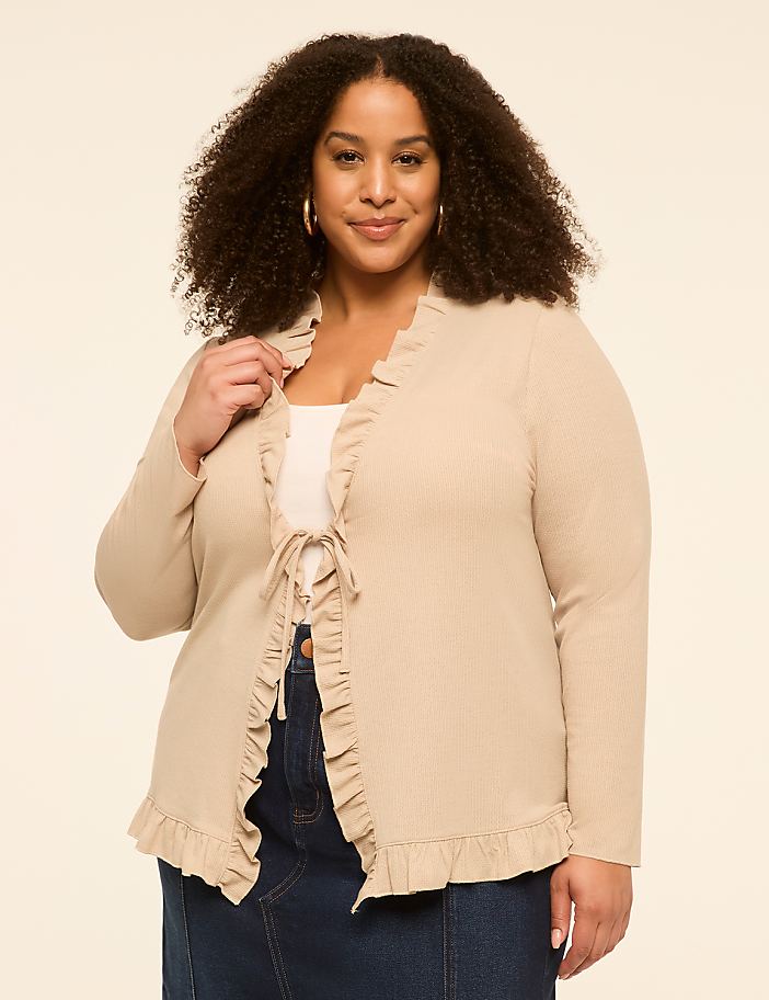 A woman with curly hair smiles at the camera, wearing a beige cardigan with ruffled trim and a white top, against a plain cream background.