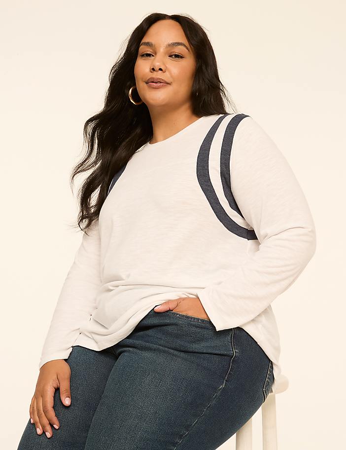 A woman with dark hair and hoop earrings poses in a white long-sleeved shirt with navy stripes on the shoulders and dark wash jeans against a plain, light-colored background.