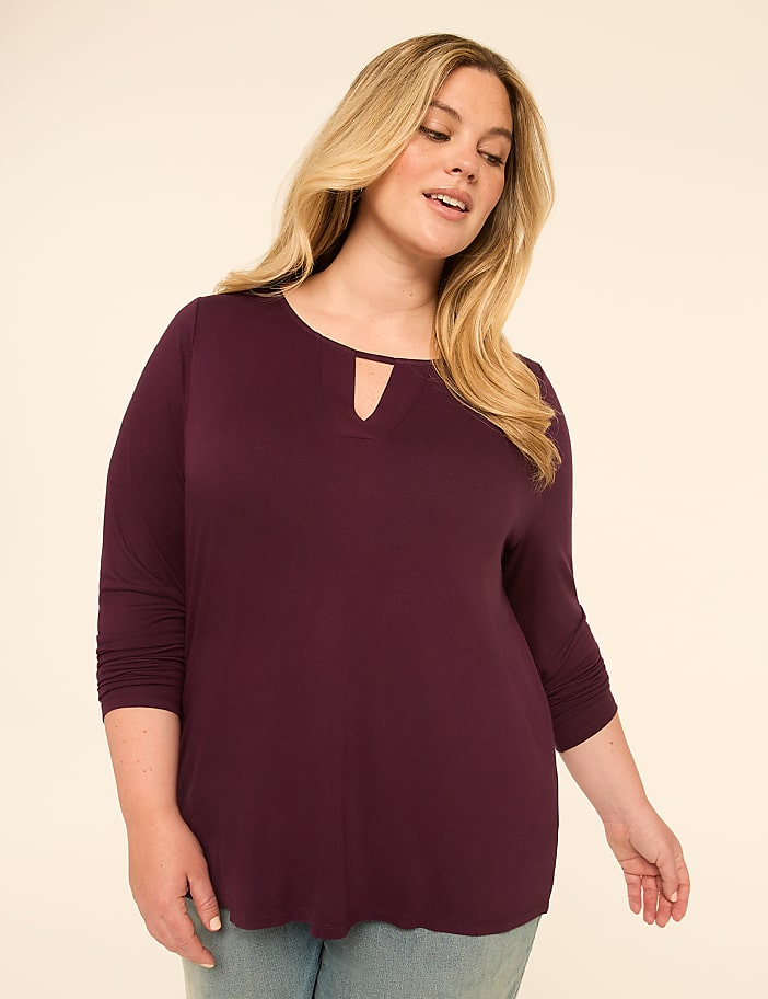 A blonde woman in a plum-colored top stands against a plain, light-colored background.