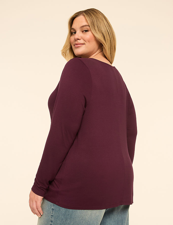 A woman in a burgundy long-sleeved shirt and jeans poses against a plain, light-colored background.