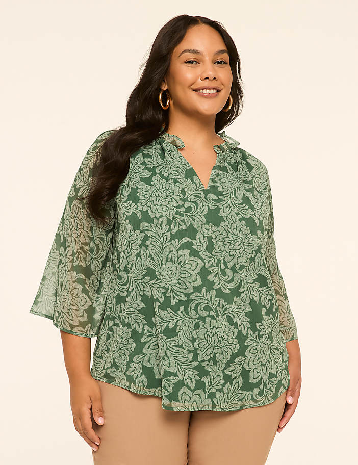 A smiling woman with long dark hair wears a green floral blouse and tan pants, standing against a plain cream background.