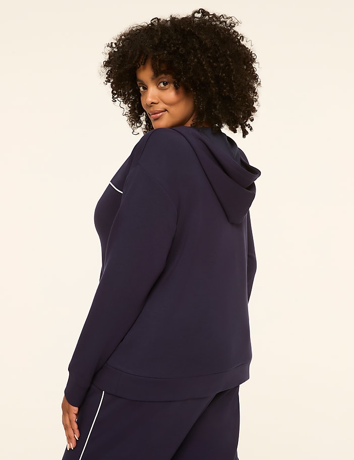A woman with curly dark hair in a navy blue hoodie and matching pants stands against a plain cream background.
