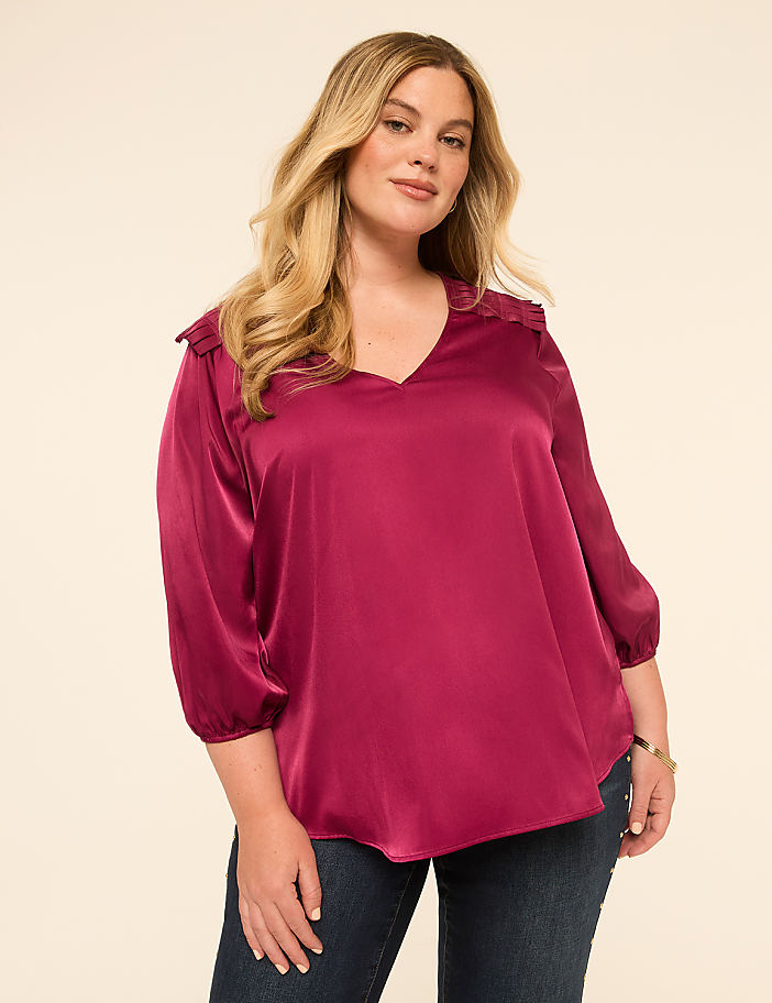 A woman in a magenta blouse and jeans stands against a plain, light-colored background.