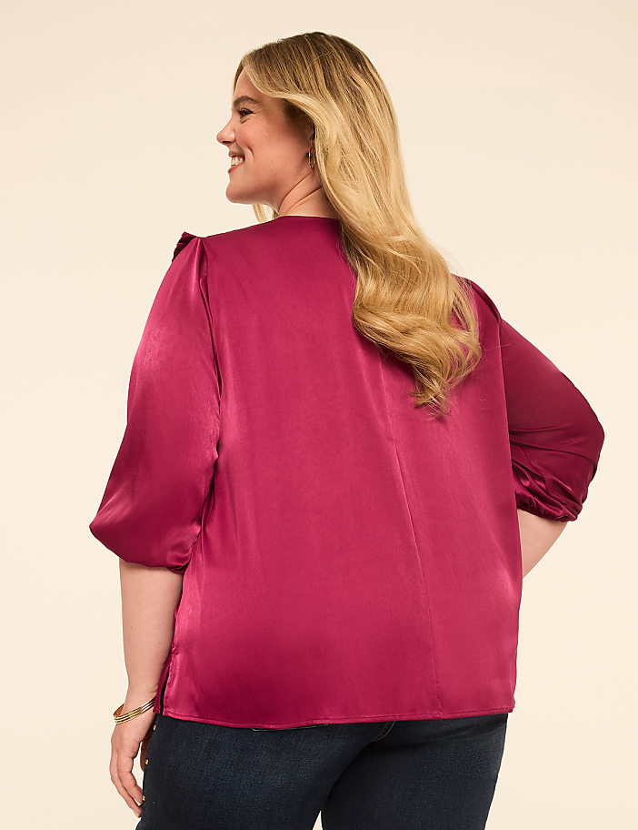 A woman in a satin magenta blouse and dark jeans stands against a plain cream background.