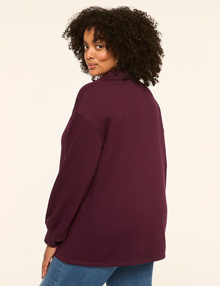 A woman with curly dark hair, wearing a burgundy turtleneck sweater and blue jeans, poses against a plain, light-colored background.