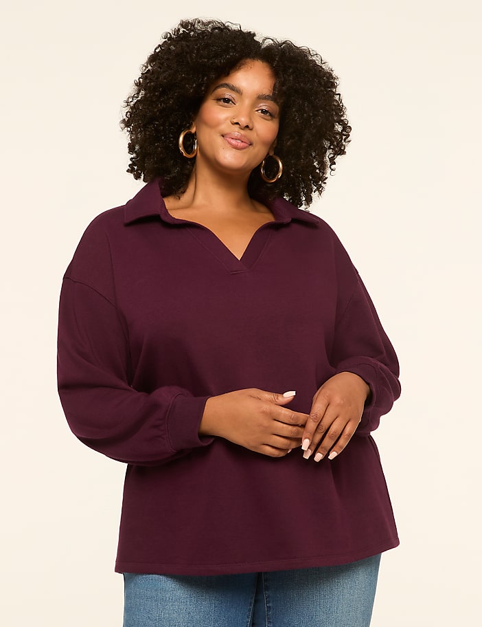 A woman with curly dark hair and hoop earrings smiles as she poses in a plum-colored collared shirt and blue jeans against a plain, light-colored background.