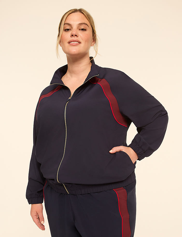 A woman in a navy blue tracksuit with maroon accents stands against a plain, light-colored background.