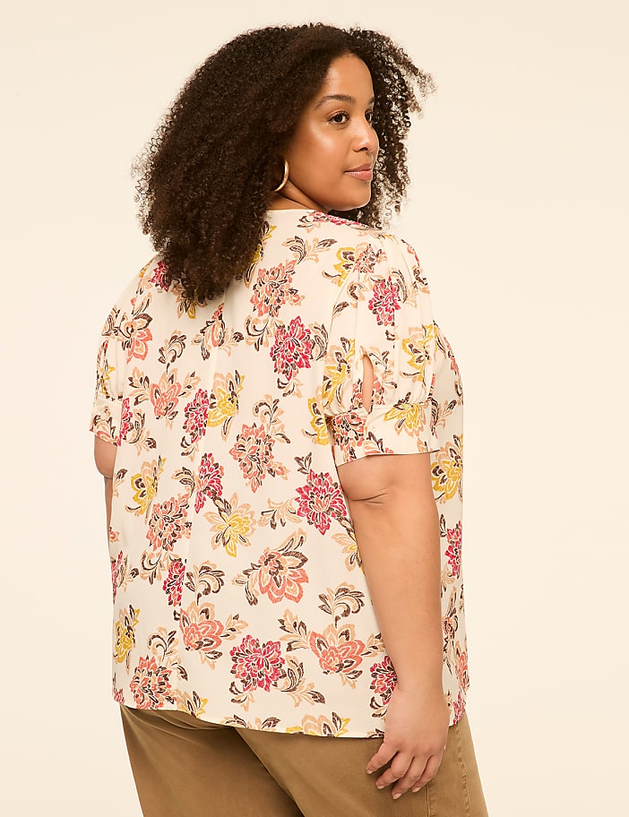 A woman with curly hair wears a floral blouse and brown pants against a plain background.