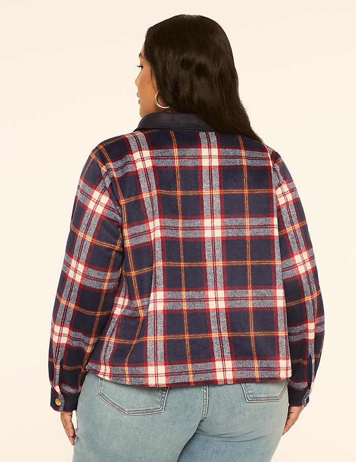 A woman in a plaid jacket and jeans stands against a plain, light-colored background.