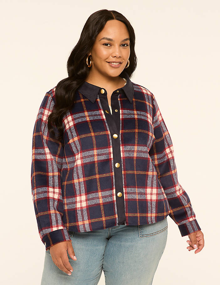 A smiling woman in a plaid jacket and jeans stands against a plain, light-colored background.