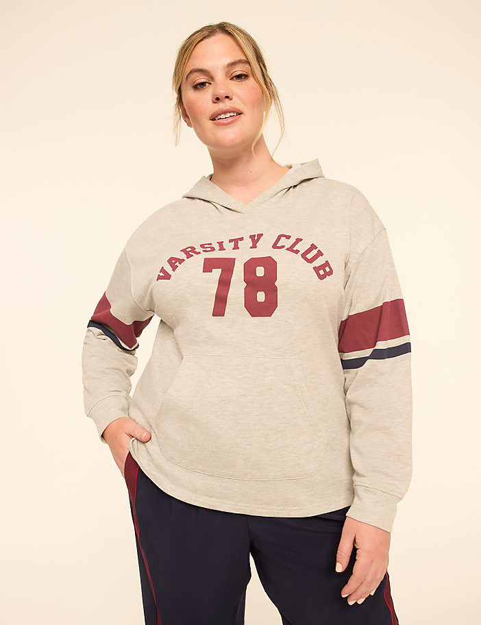 A woman in a grey "Varsity Club 78" hoodie and navy pants stands against a plain beige background.