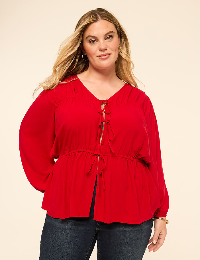 A woman in a red blouse and jeans stands against a plain, light-colored background.