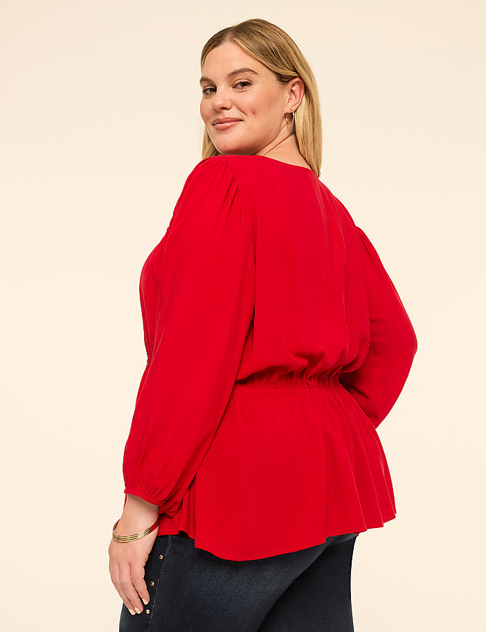 A woman in a red peplum top and dark jeans poses against a plain, light-colored background.