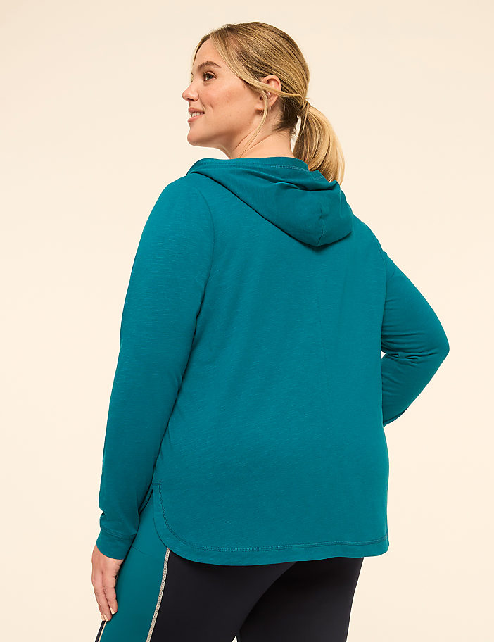 A woman in a teal hoodie and dark leggings stands against a plain, light-colored background.