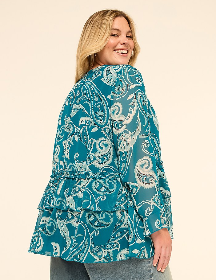 A woman in a teal paisley blouse with ruffled tiers smiles over her shoulder against a plain cream background.