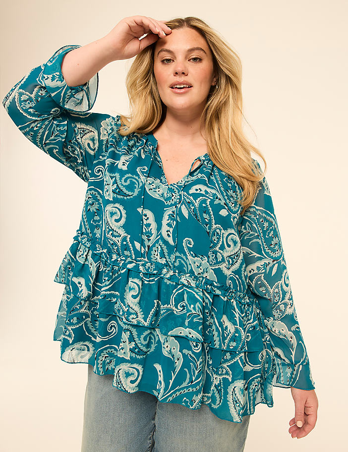 A woman in a teal paisley-print ruffled blouse and jeans poses against a plain beige background.