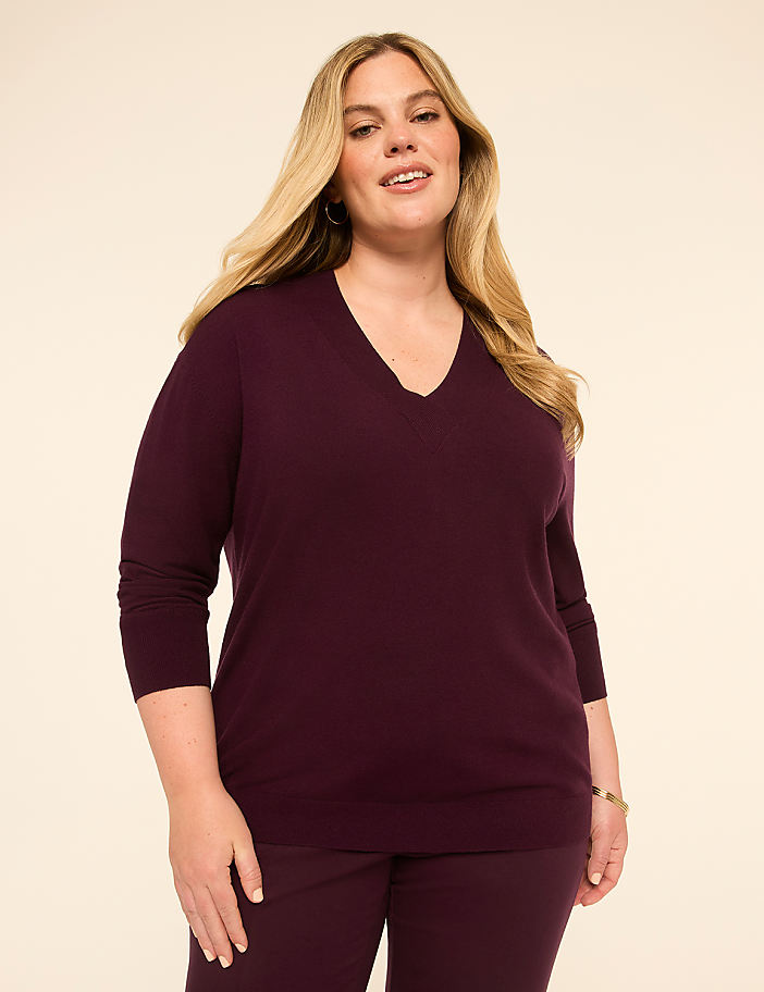 A woman in a plum v-neck sweater and matching pants stands against a plain, light beige background.