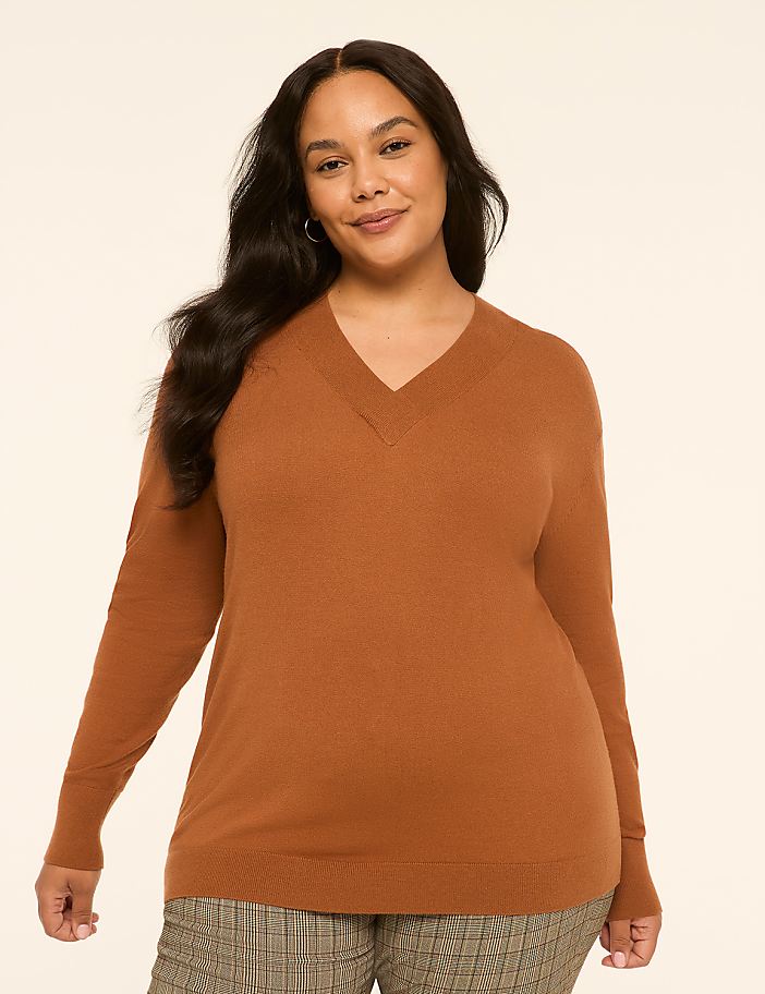 A woman in a brown v-neck sweater and plaid pants stands against a plain, light-colored background.