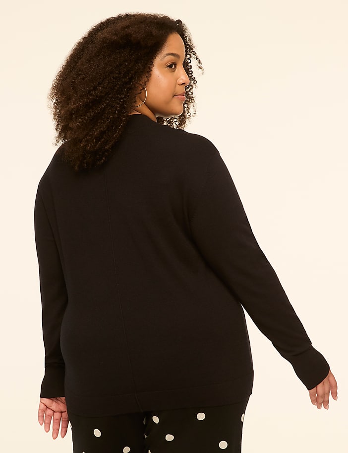 A woman with curly dark hair, wearing a black sweater and polka dot pants, stands against a plain, light-colored background.