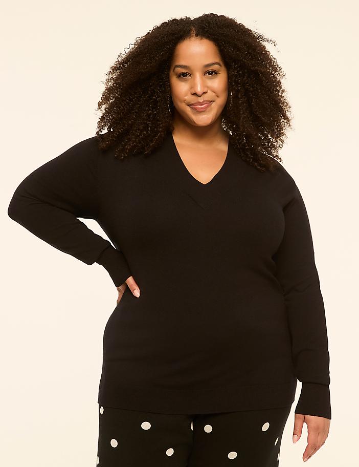 A woman with curly hair and a black v-neck sweater stands against a plain, light-colored background, her lower body adorned with polka-dot pants.