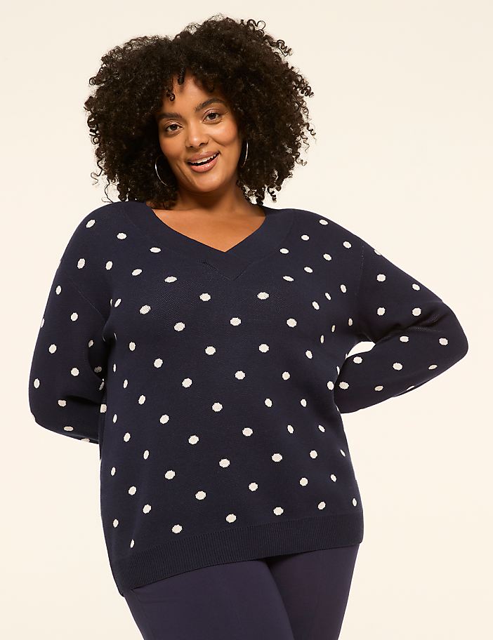 A smiling woman in a navy polka dot sweater stands against a plain, light-colored background.