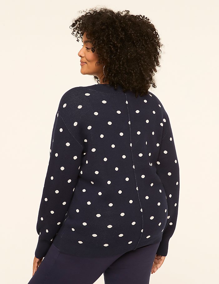 A woman in a navy polka dot sweater stands against a plain beige background.