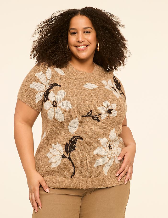 A smiling woman with curly hair wears a tan, floral-patterned sweater and tan pants against a plain, light tan background.