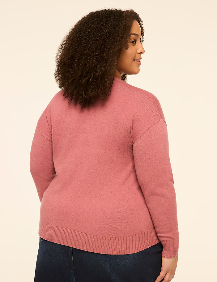 A woman in a pink sweater and dark skirt stands against a plain, light-colored background.