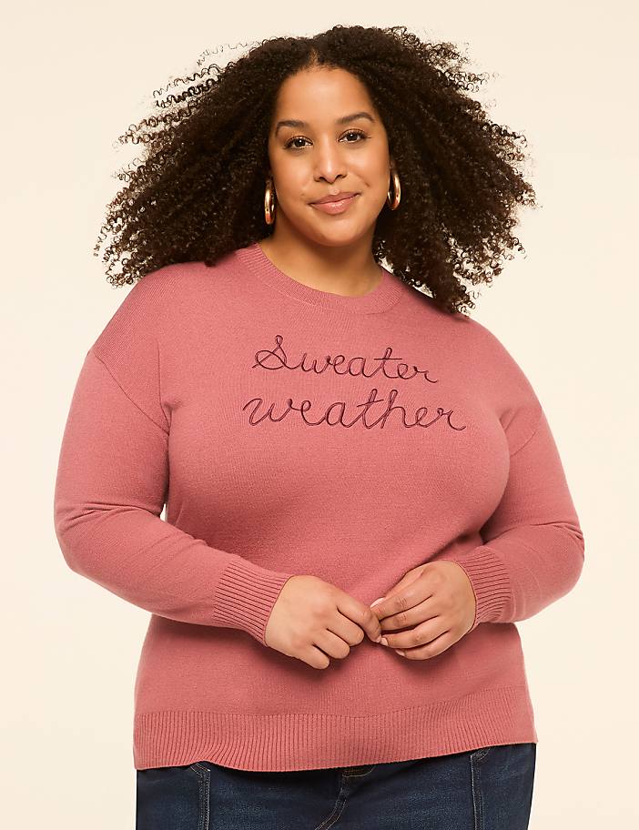 A woman with curly hair smiles in a pink "Sweater weather" sweatshirt against a plain, light-colored background.