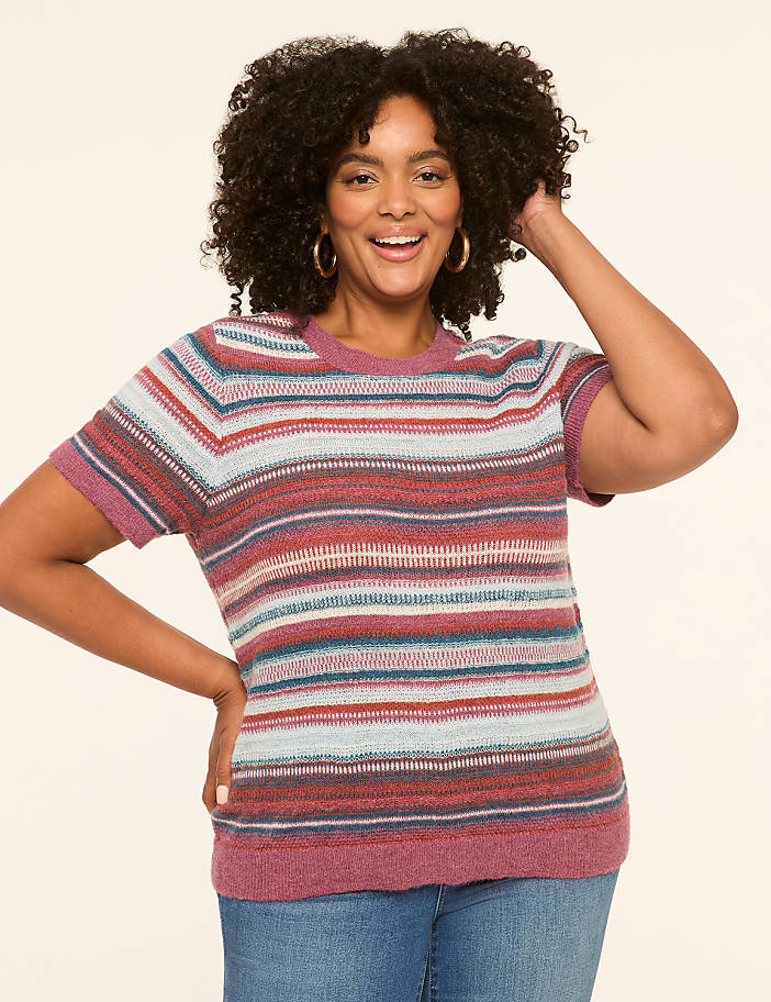 A smiling woman with curly hair in a striped sweater and jeans poses against a plain, light-colored background.