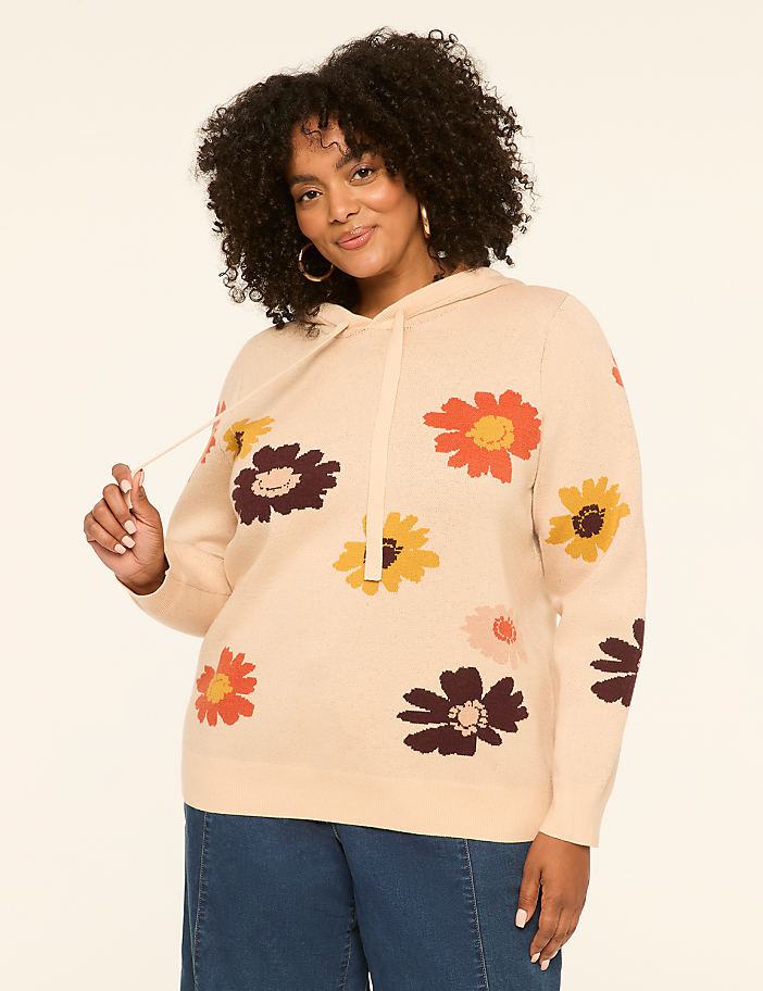 A woman in a cream-colored hoodie with a floral pattern stands against a plain, light background.
