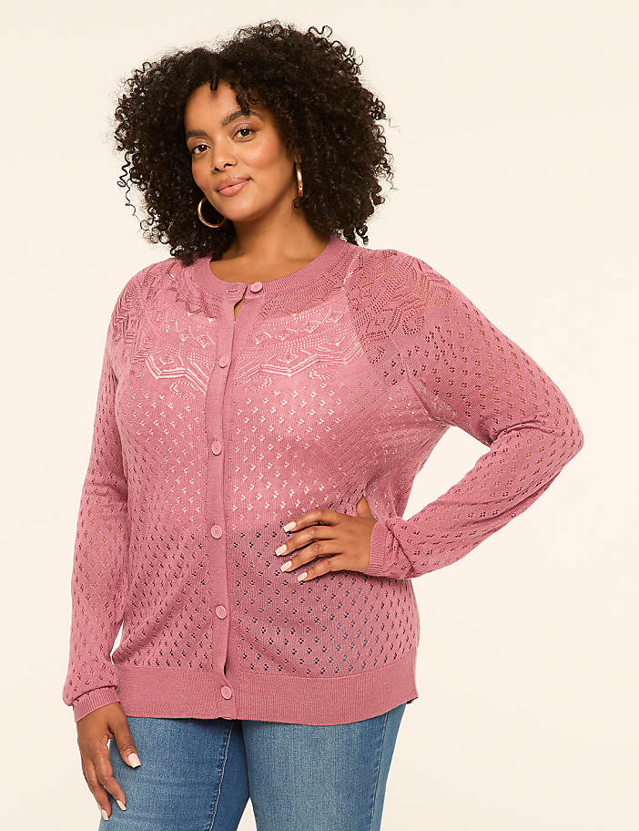 A woman in a pink cardigan and jeans stands against a plain, light-colored background.