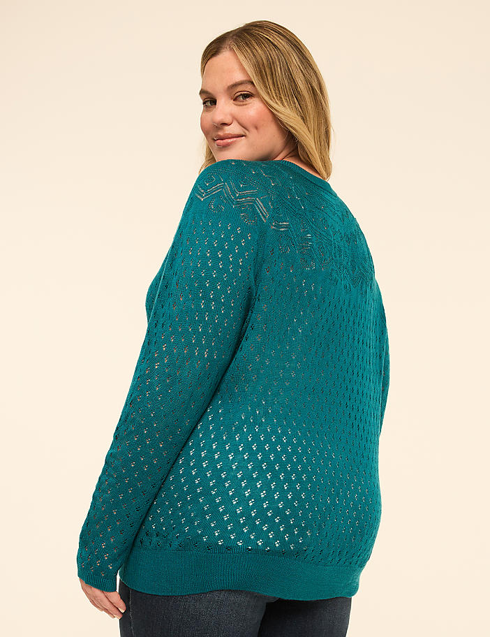 A woman in a teal, open-knit sweater turns to smile over her shoulder against a plain, light-colored background.