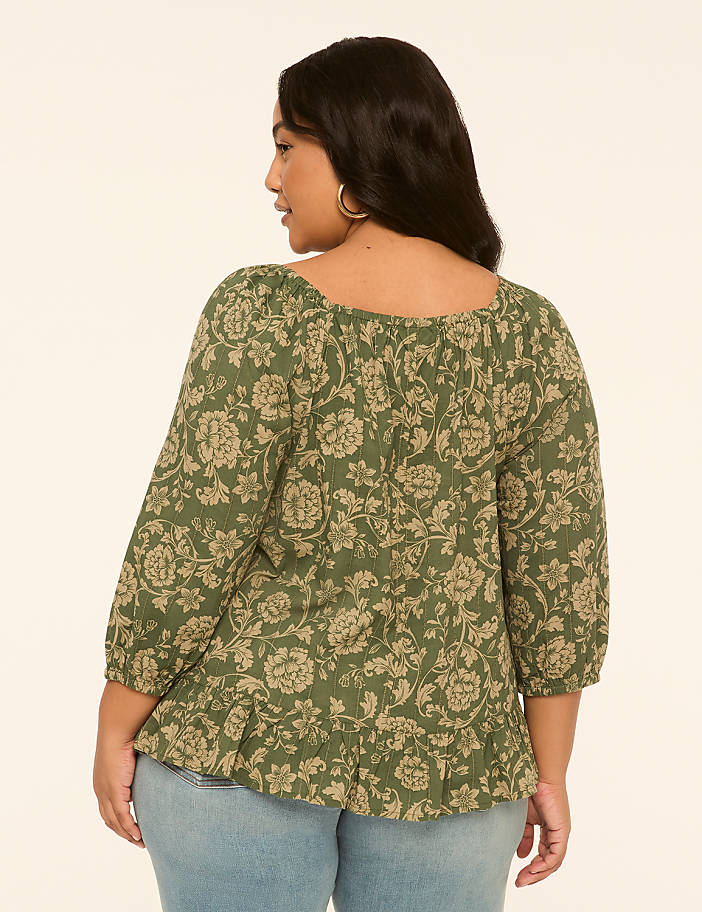 A woman in a floral green top and blue jeans stands against a plain, light-colored background.
