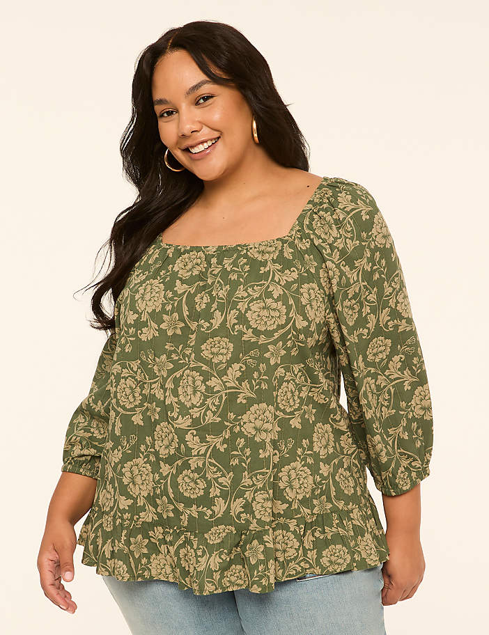 A smiling woman with dark hair and hoop earrings wears a green floral-print peasant blouse with a square neckline and light blue jeans, set against a plain cream background.