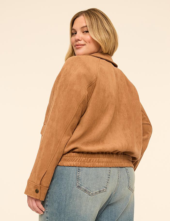 A woman in a tan jacket and blue jeans poses against a plain, light-colored background.