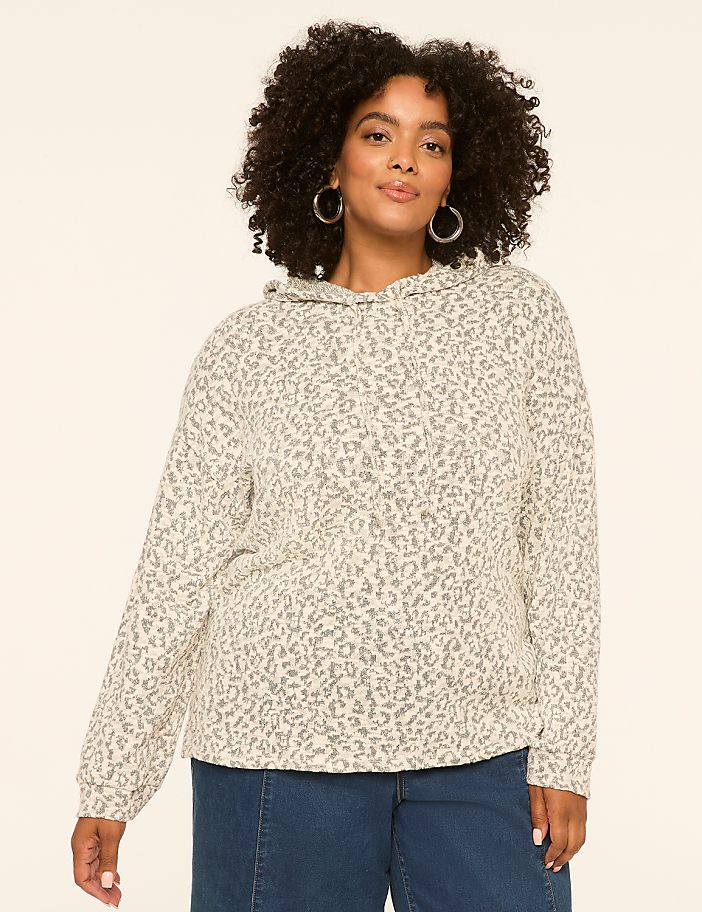 A woman with curly dark hair and hoop earrings stands in the foreground wearing a leopard-print hoodie and blue jeans, against a plain, light-colored background.