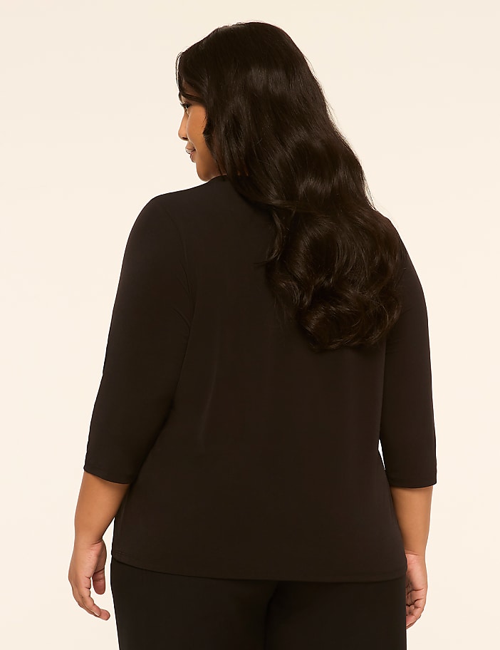 A woman with long, dark hair stands facing away from the camera in a black top and pants against a plain, light-colored background.