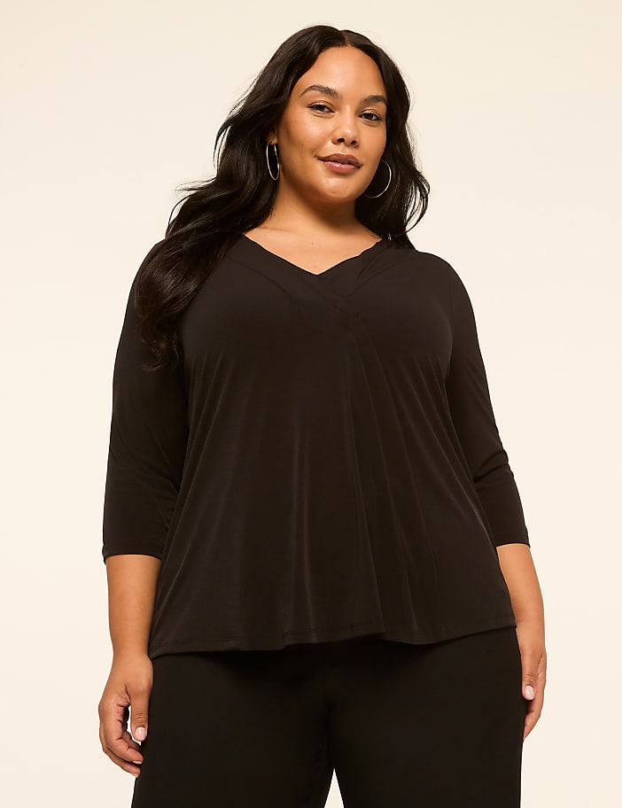 A woman with dark hair and hoop earrings poses in a black v-neck top and black pants against a plain beige background.