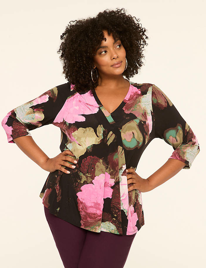 A woman with curly dark hair and hoop earrings poses with her hands on her hips, wearing a black top with a colorful abstract floral print and dark purple pants, against a plain cream-colored background.