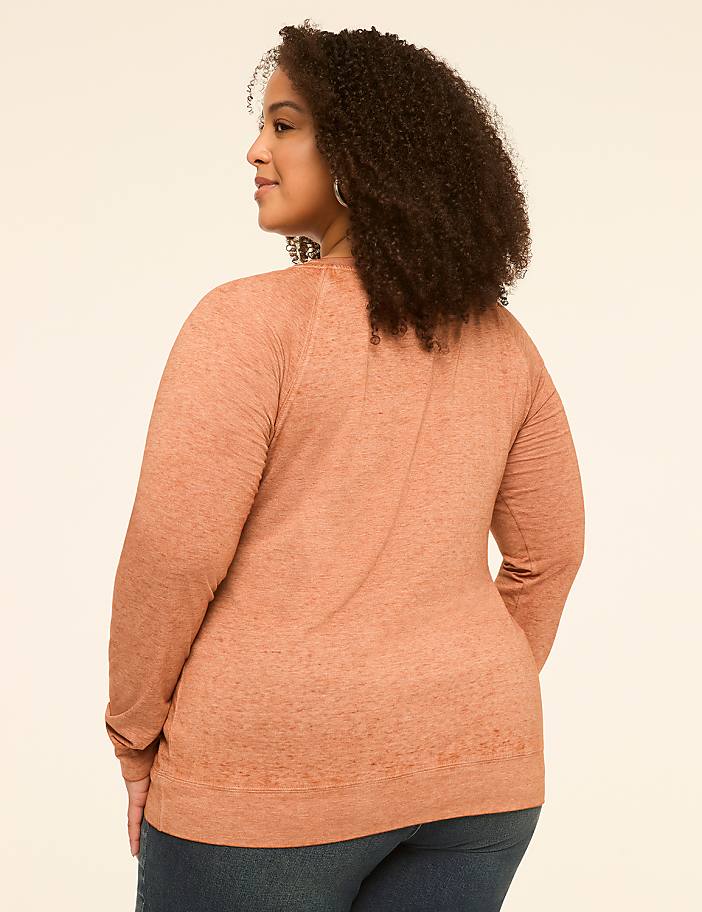 A woman with curly brown hair, wearing a peach-colored long-sleeved shirt and blue jeans, stands against a plain, light-colored background.