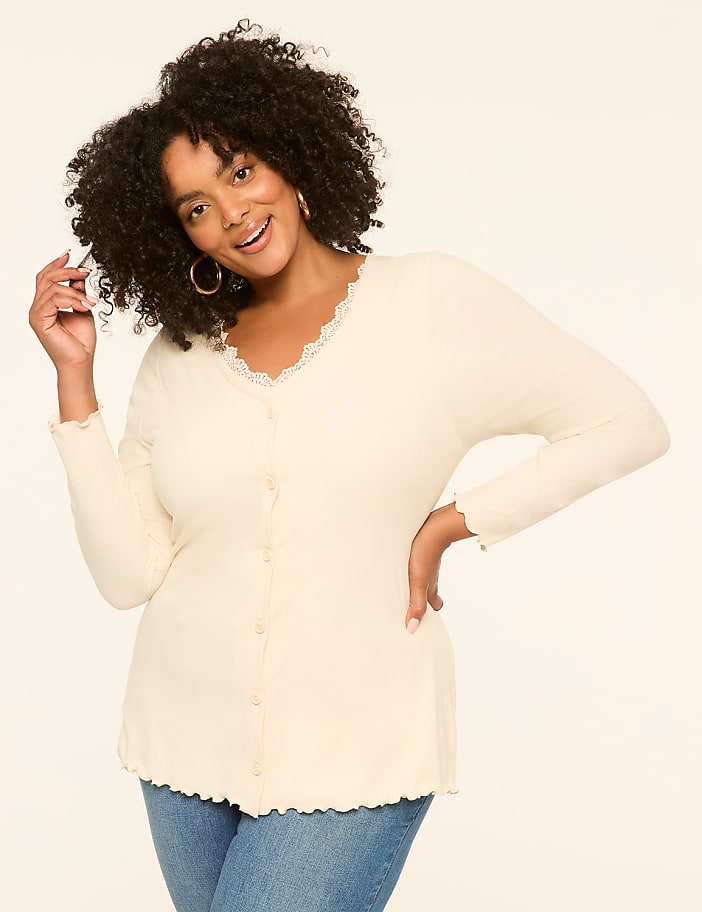 A smiling woman with curly hair in a cream-colored top and blue jeans poses against a plain, light-colored background.