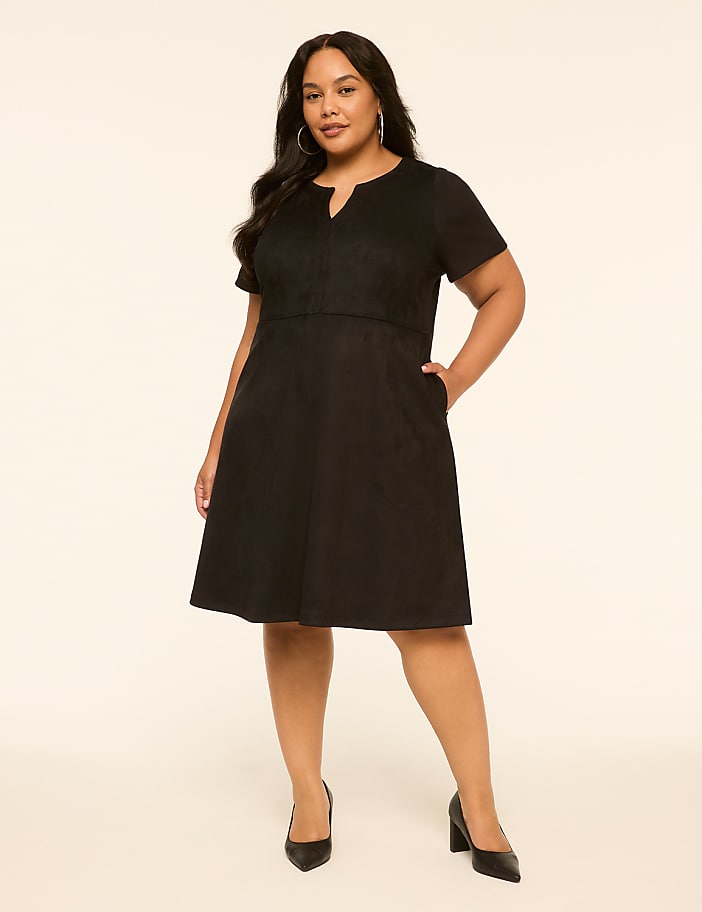 A plus-size woman in a black dress and heels stands against a plain, light-colored background.