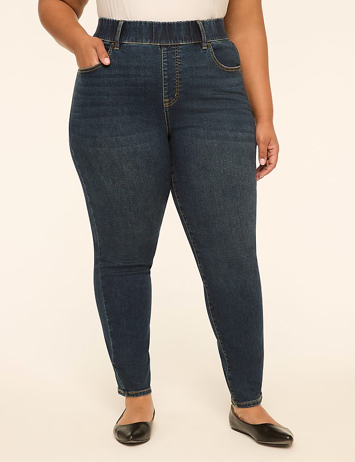 A woman in dark wash denim jeans and black ballet flats stands against a plain, light-colored background.