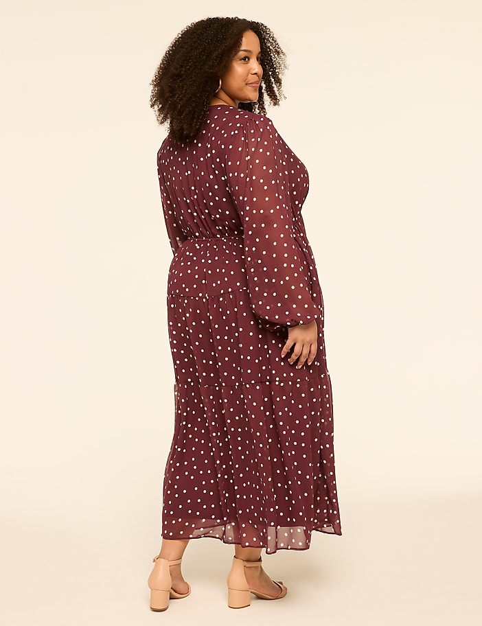 A woman in a burgundy polka dot dress stands against a plain, light-colored background.