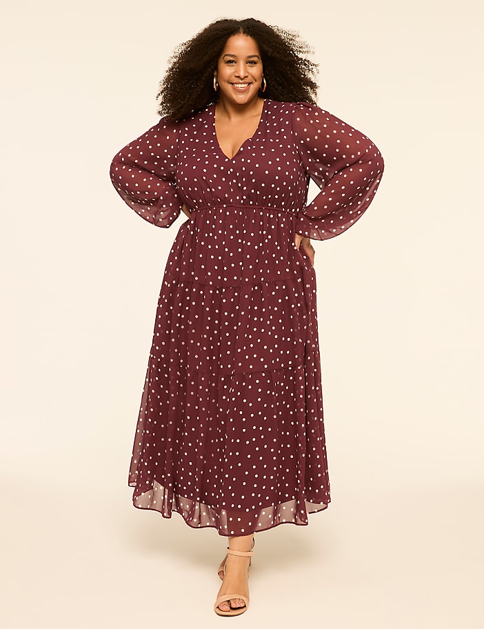 A smiling woman with curly hair stands in a maroon polka-dot dress and beige heels against a plain cream background.