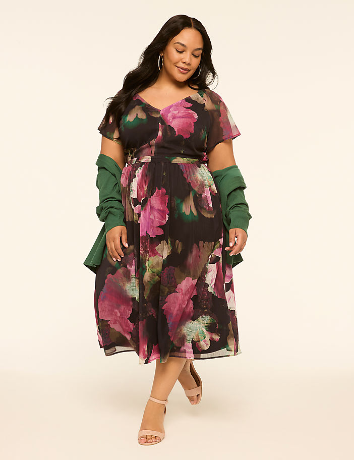 A woman in a floral dress and green jacket stands against a plain, light-colored background.