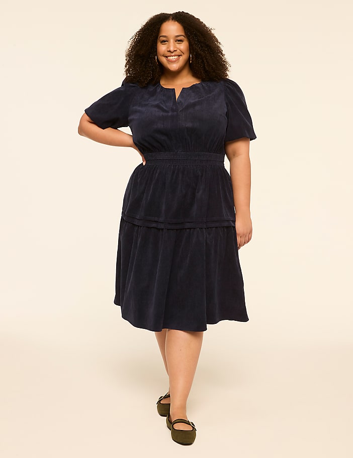 A smiling woman in a navy blue dress stands against a plain, light-colored background.